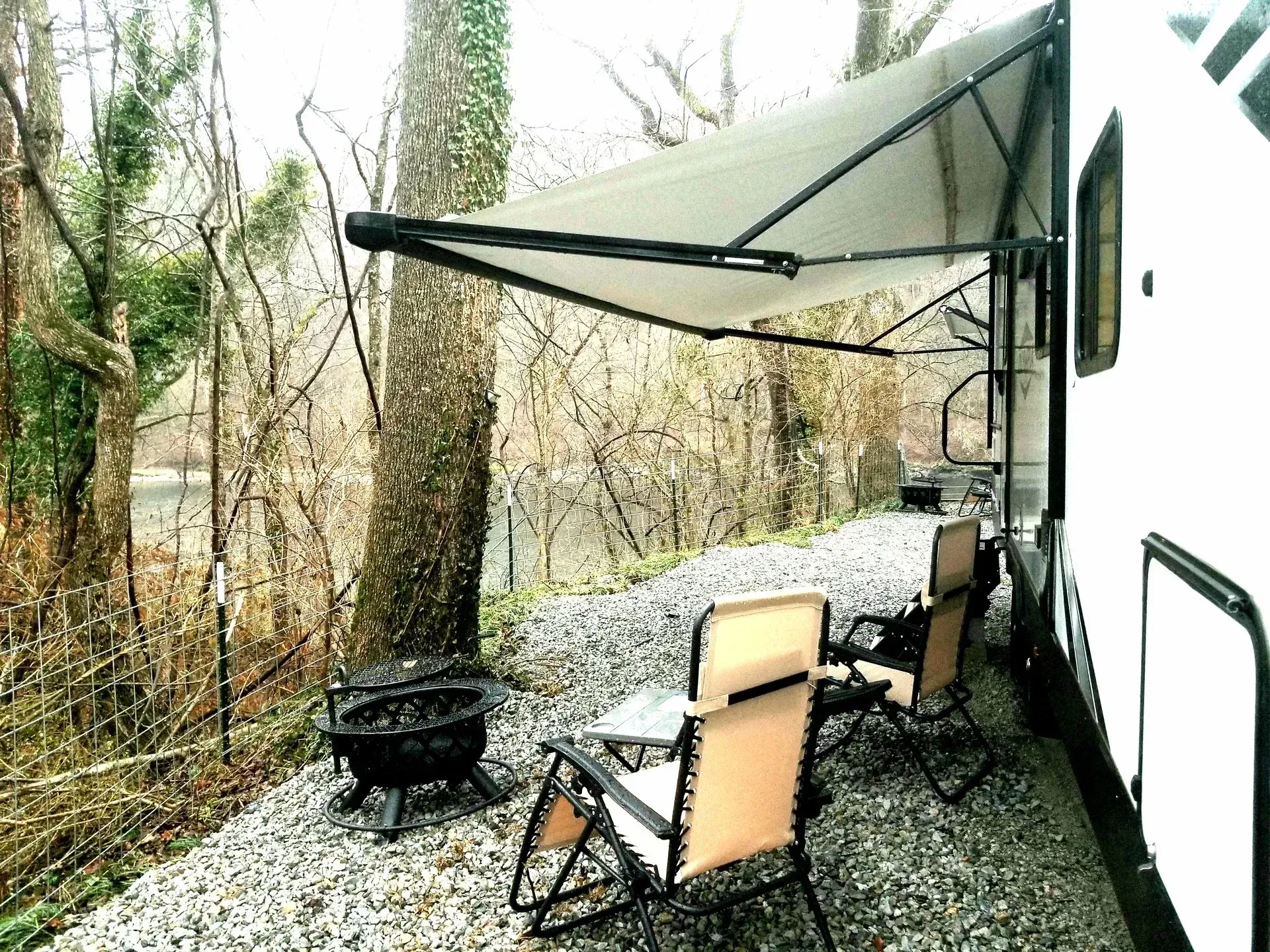 A rv with a table and chairs under an awning in the woods.