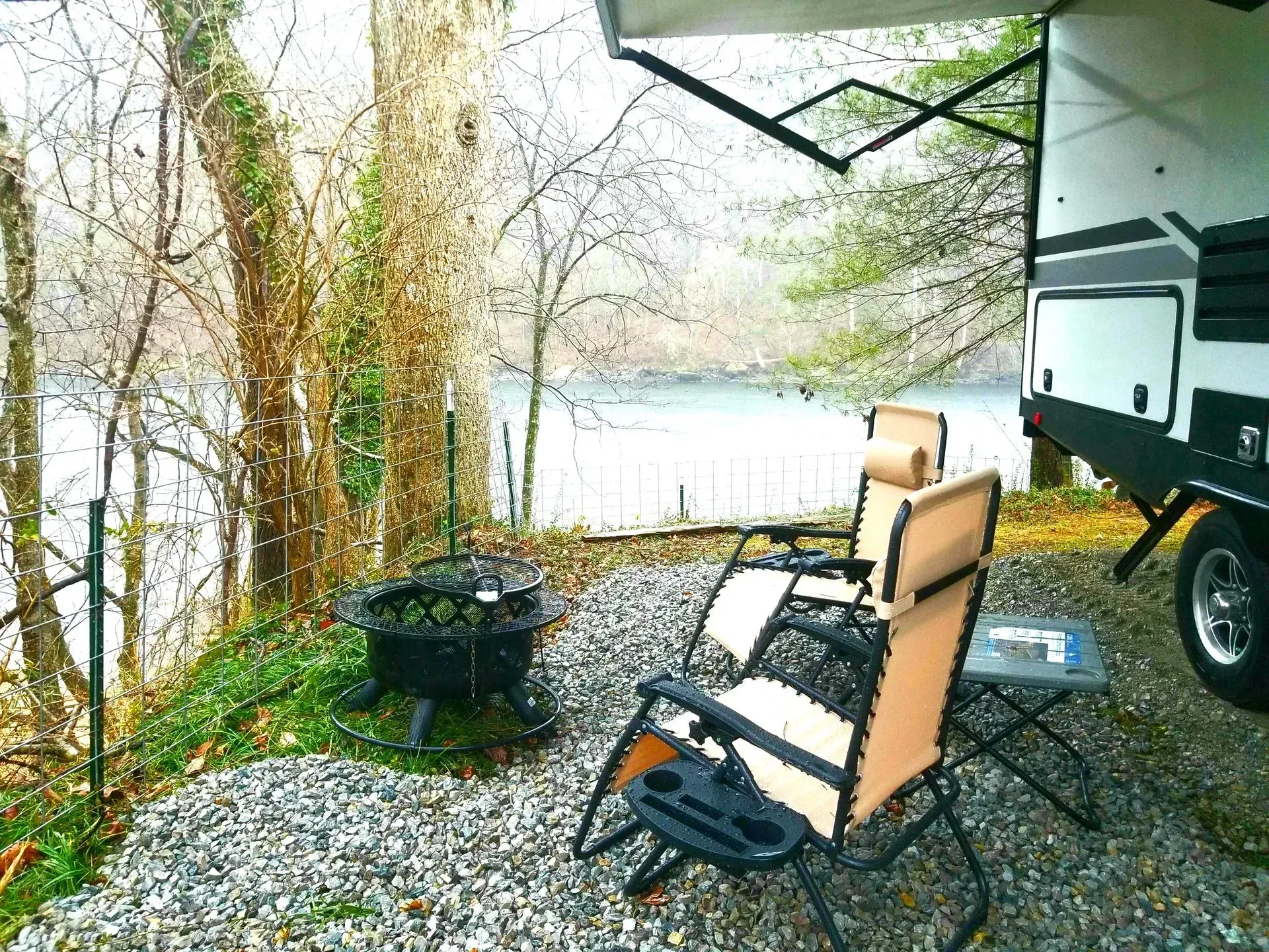 A rv parked next to a lake with chairs and a fire pit.