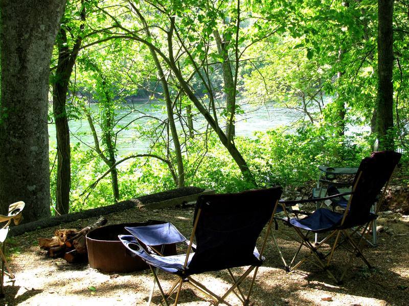 Two folding chairs are sitting next to a river in the woods.