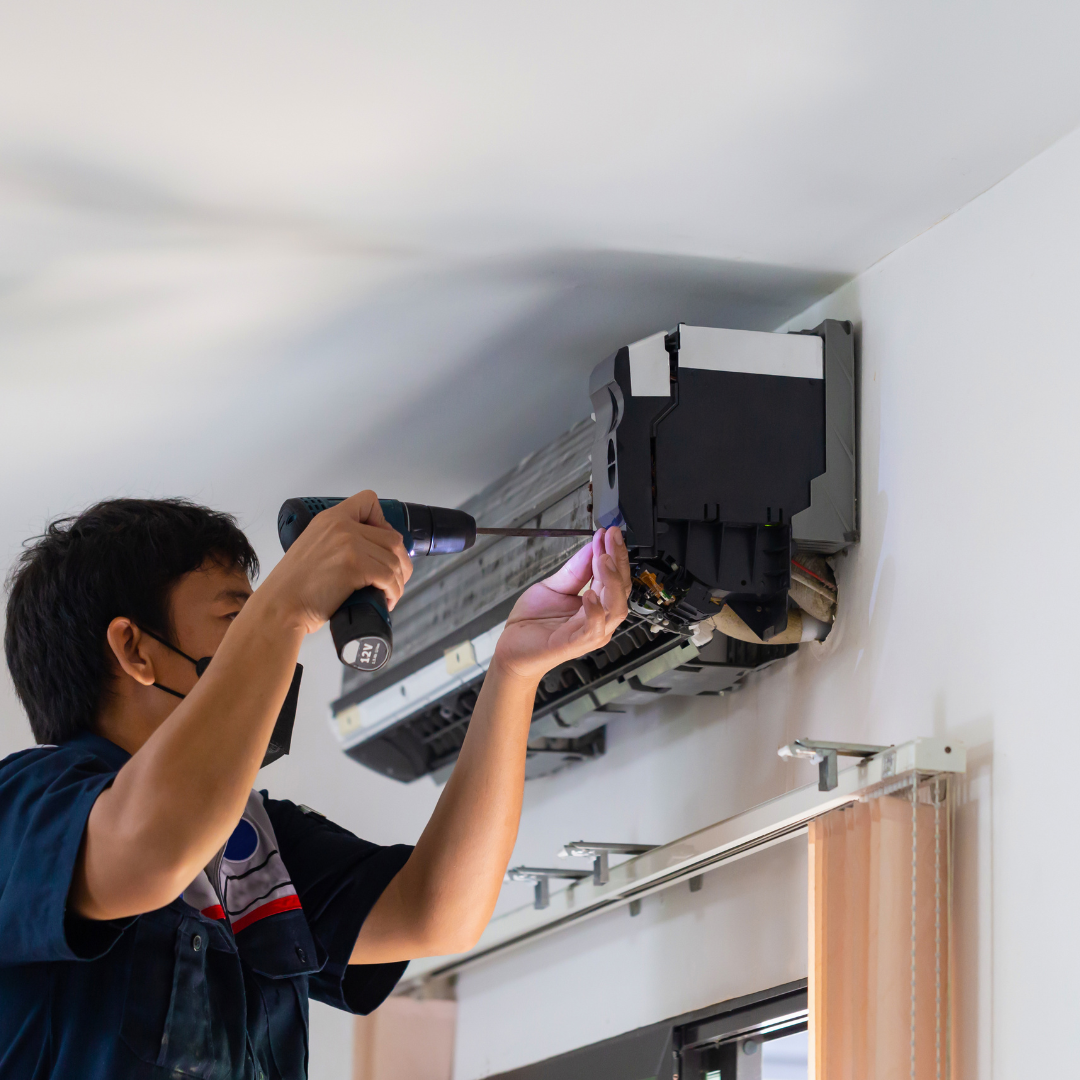 Asian technician in mask repairs wall-mounted air conditioner with a power drill.