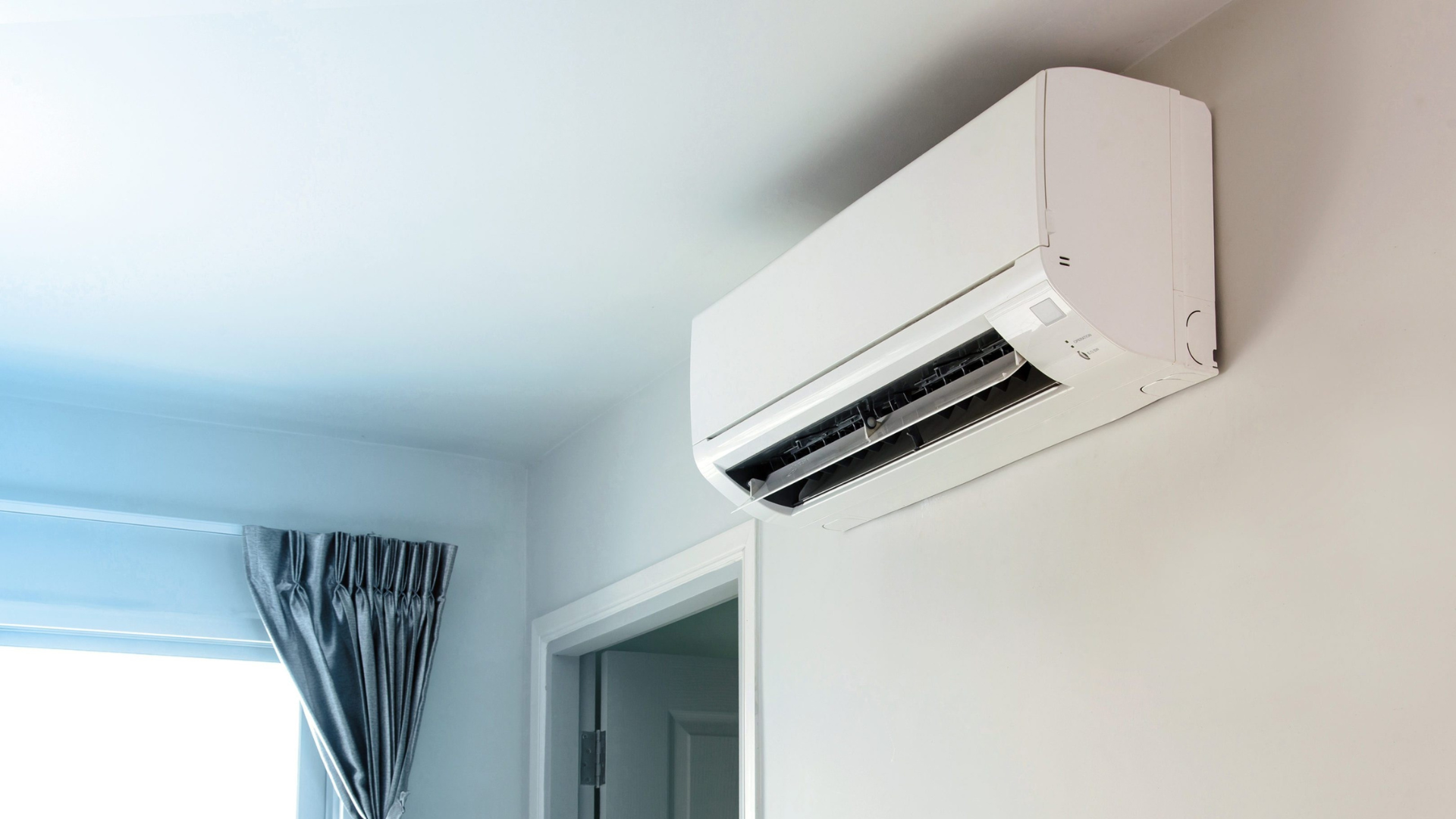 White air conditioner mounted on a light-colored wall in a room with a window and door.