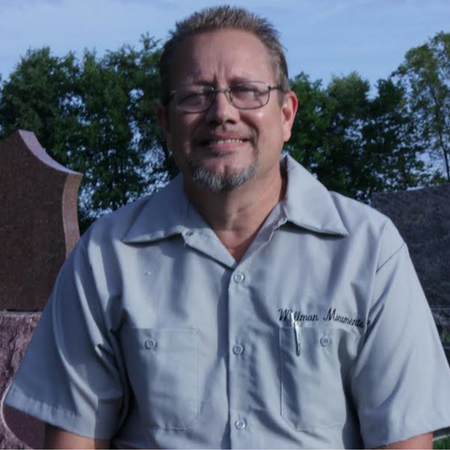 Smiling man in glasses and a light gray shirt standing outdoors with trees and a stone monument behind him