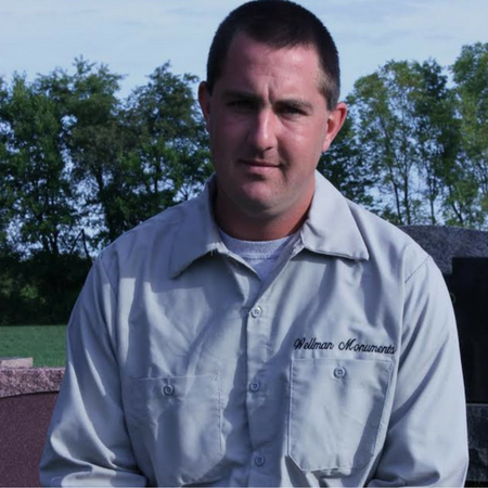 Man in gray work shirt standing outdoors in front of trees and a building.