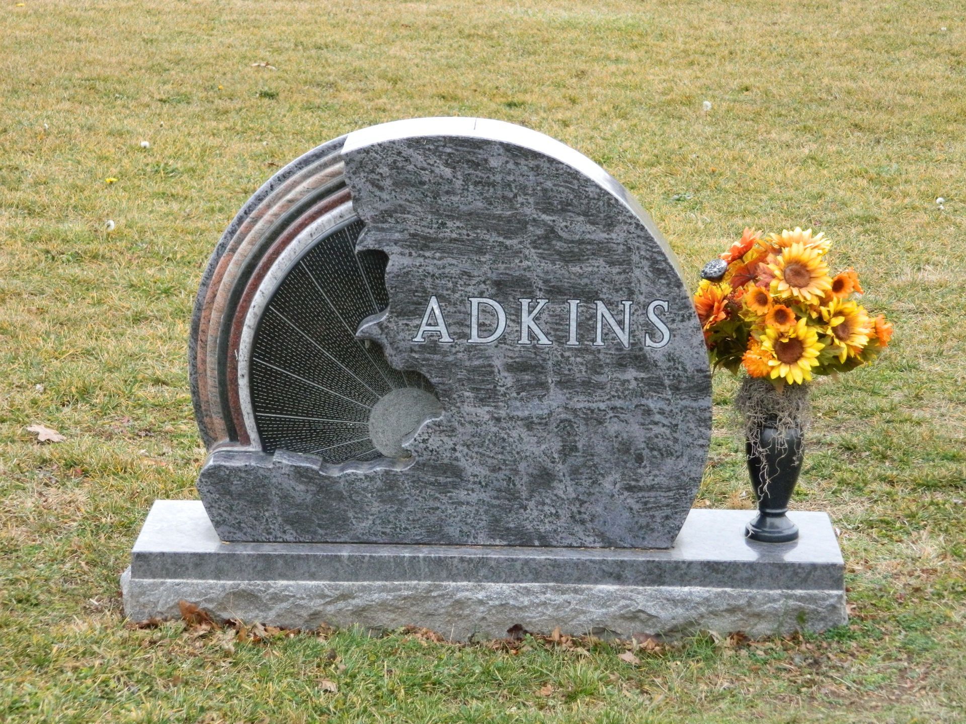 Granite gravestone engraved “ADKINS” with a vase of yellow and orange flowers on grass.