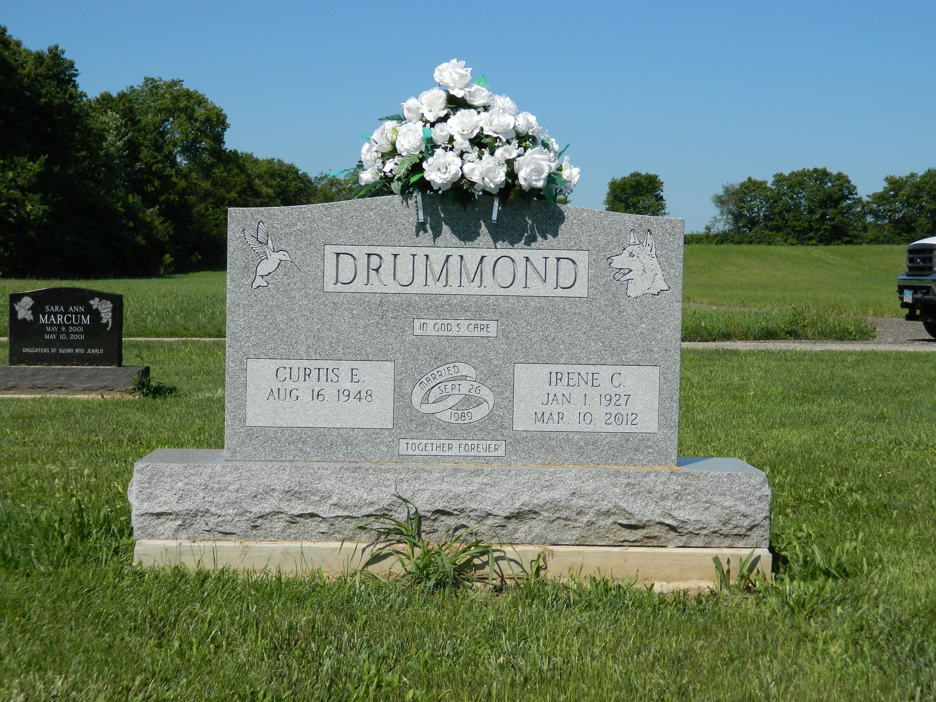 Gravestone marked DRUMMOND with white flowers in a sunny cemetery field.
