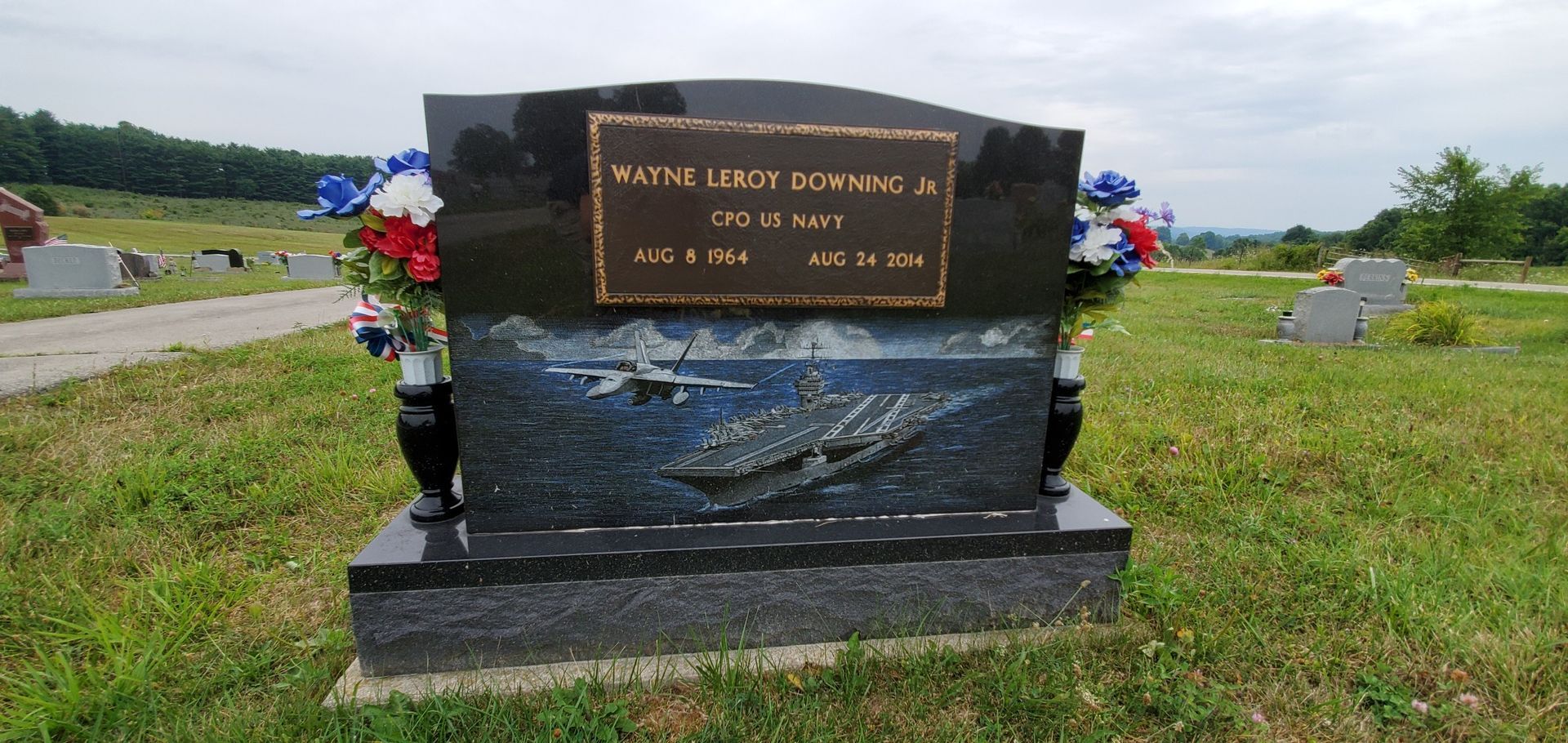 Memorial gravestone with engraved plaque and floral tributes in a grassy roadside setting