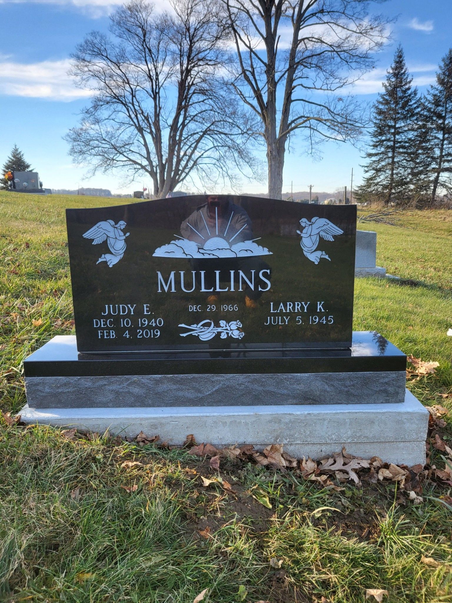 Black Mullins gravestone with angel carvings in a grassy cemetery under a blue sky