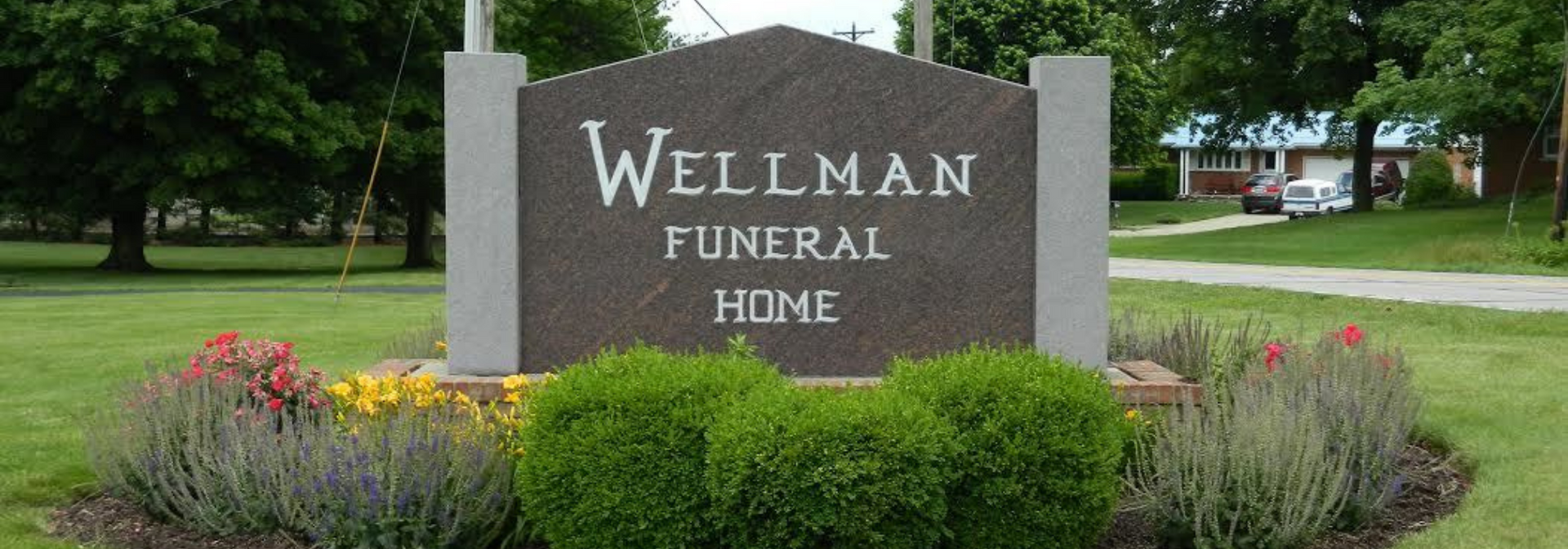 Stone sign for Wellman Funeral Home with flowers and shrubs in front.