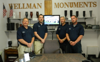 Four employees stand in a Wellman Monuments showroom beneath company signage and display shelves.