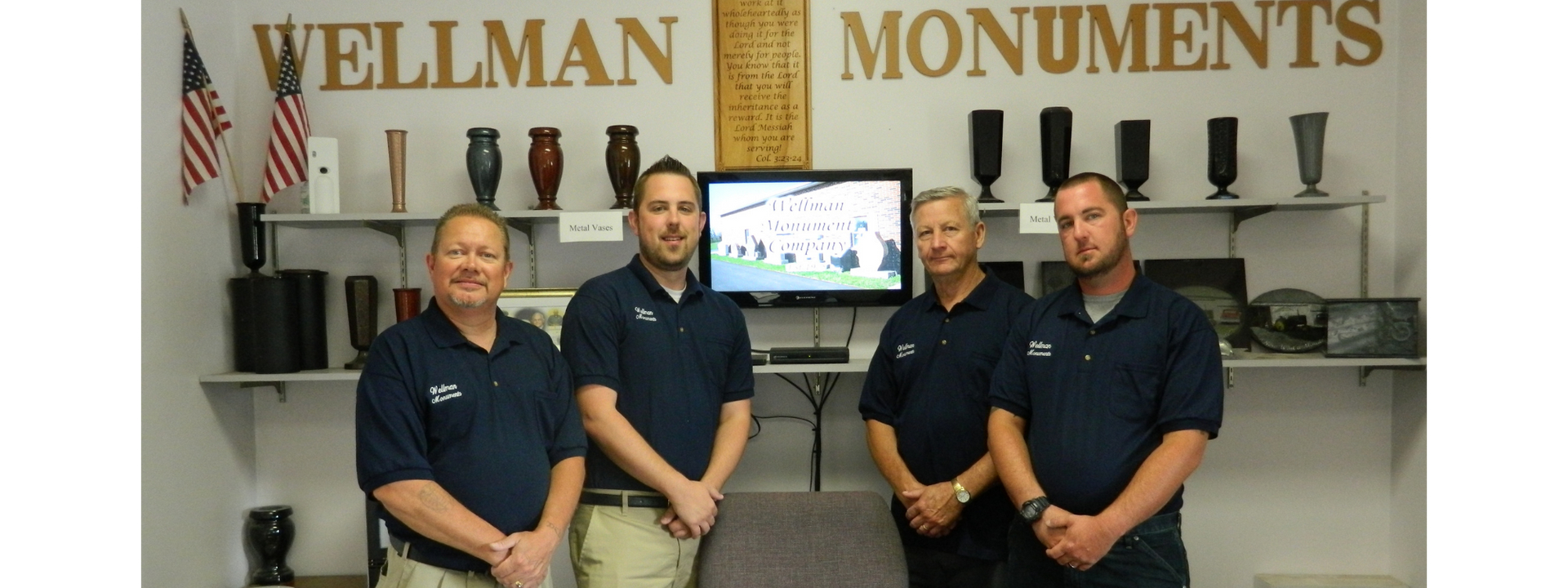 Four people standing in a monument showroom with a display monitor and large “WELLMAN MONUMENTS” sign behind them