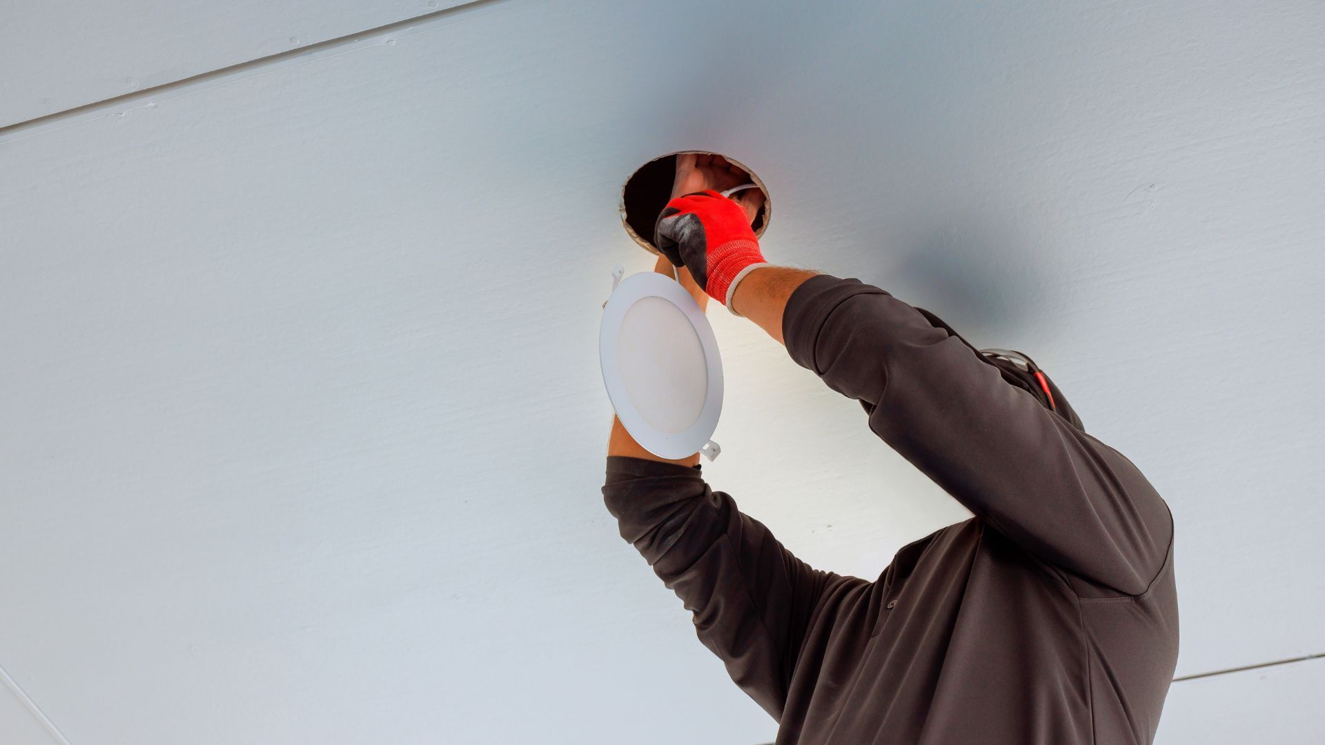 A person wearing work gloves installing a circular recessed light fixture into a white ceiling.