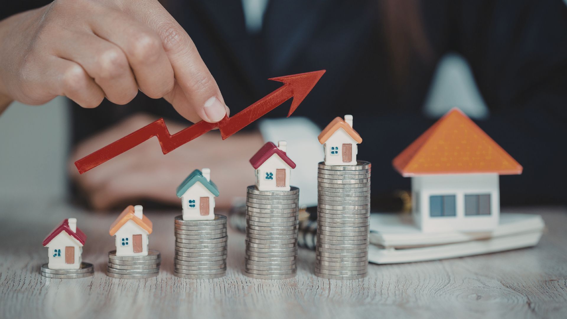 A hand points toward a red arrow rising above four stacks of coins topped with miniature houses, indicating growth.