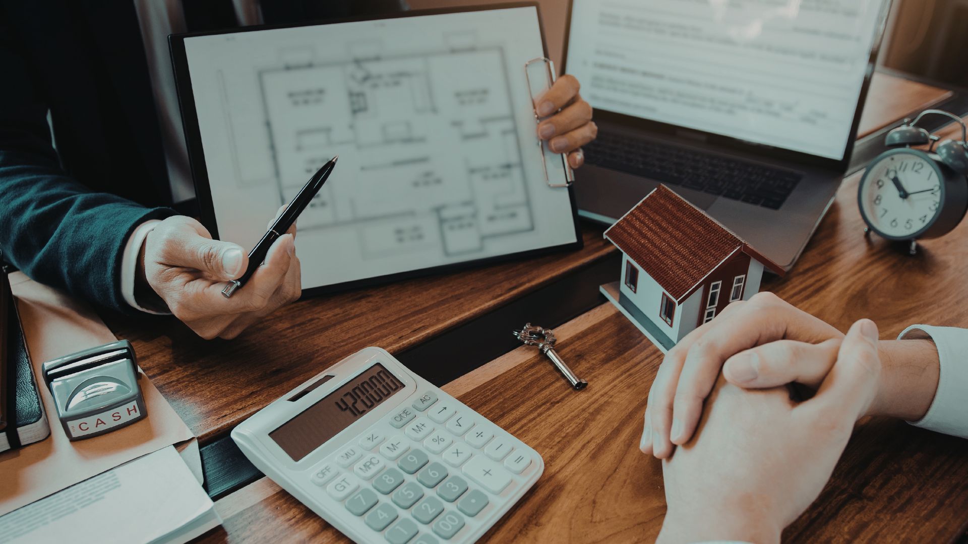 Two people sit at a wooden desk with a floor plan, calculator, and house model, discussing real estate or property plans.