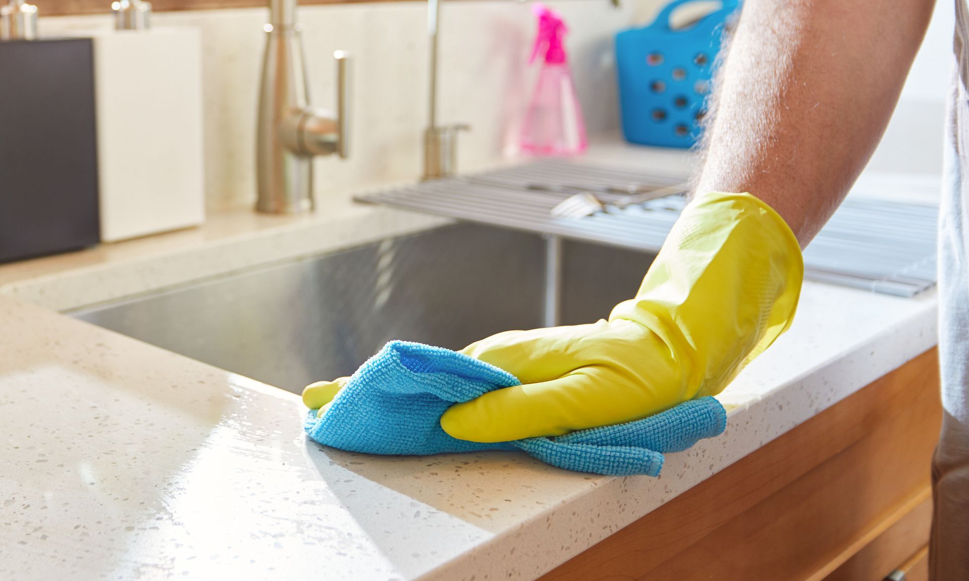 Hand in yellow glove wiping a kitchen countertop with a blue cloth near a sink.