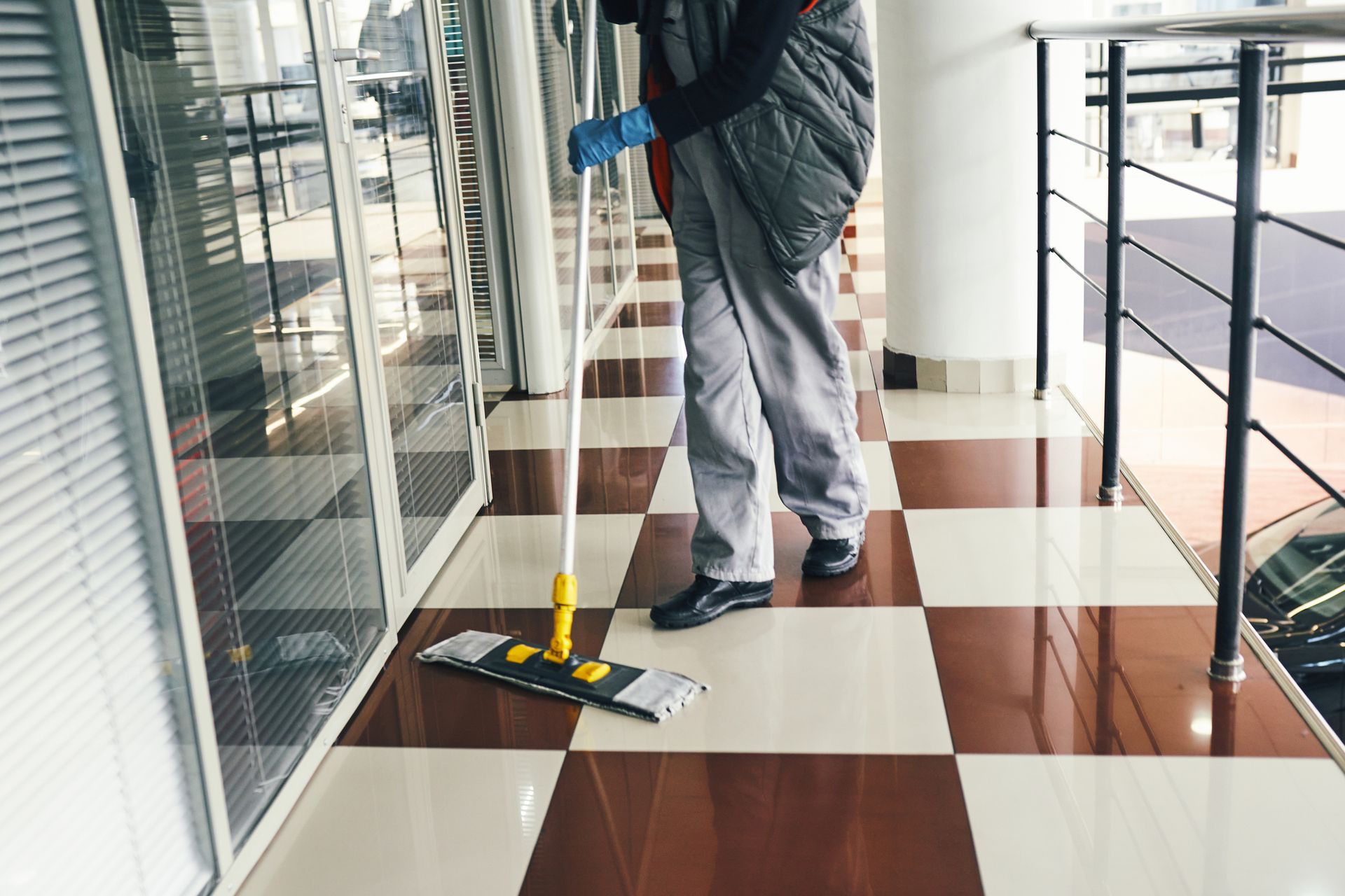 Person mopping a checkered floor in a hallway with windows and a railing.