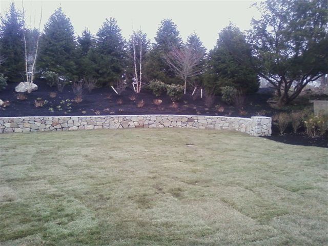 Curved stone retaining wall beside a grassy lawn, with trees and shrubs along the slope.