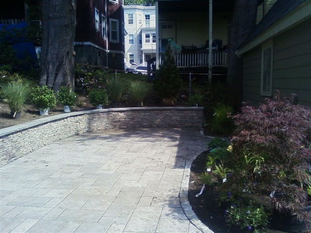Paved courtyard path with raised stone wall, flowers, and greenery between buildings