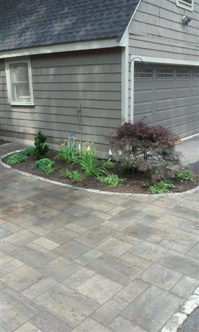 Driveway beside a gray house with a small landscaped garden bed and paver stone paving.