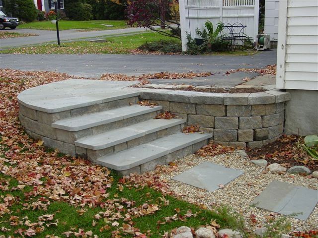Concrete steps leading to a raised patio beside a house, with stone retaining wall and fallen leaves nearby