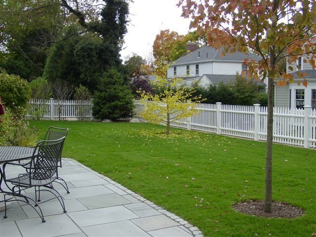 Backyard patio with metal table and chairs, green lawn, white fence, and autumn trees