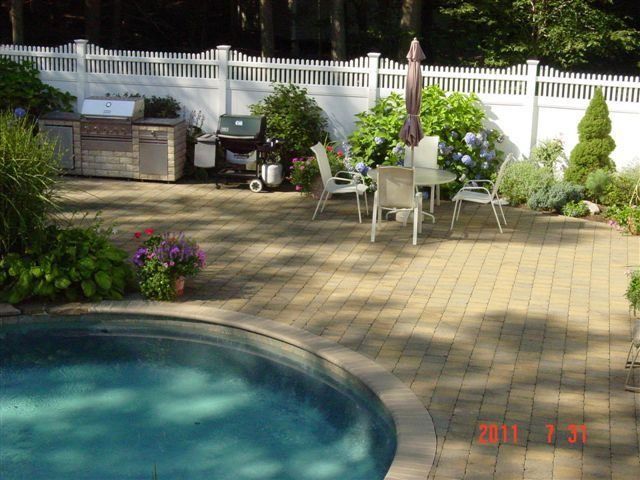 Backyard patio with a swimming pool, white chairs and table, grill, and potted plants in bright sun