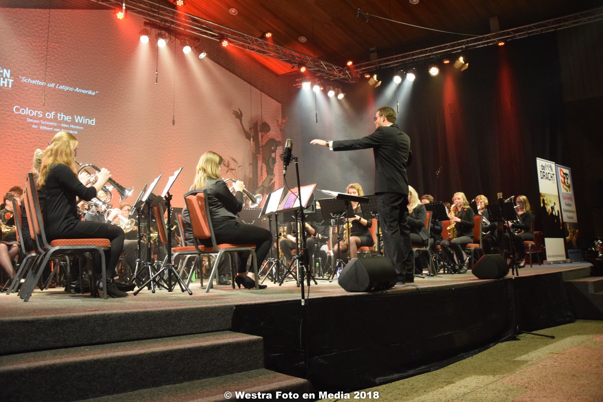 Orkest treedt op op een podium onder rood en wit licht, de dirigent leidt de musici in galakleding.