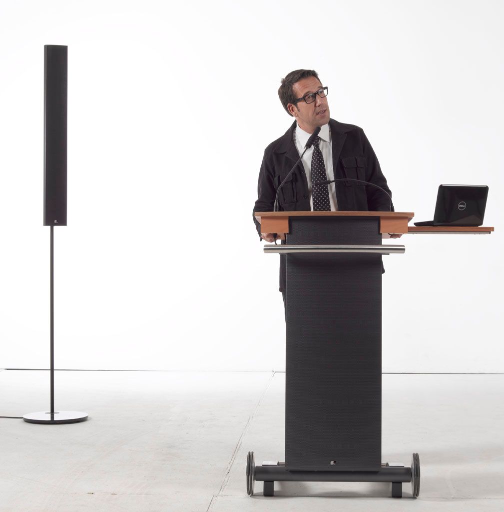 Een man staat achter een podium te spreken, naast een laptop, op een minimalistisch wit podium.
