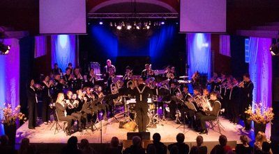 Orkest en koor treden op een verlicht podium op met paarsblauwe verlichting, met een dirigent in het midden vooraan.