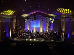 Kerstconcert in een versierde kerkzaal met koor, orkest, verlichte kerstboom en paarse podiumverlichting.
