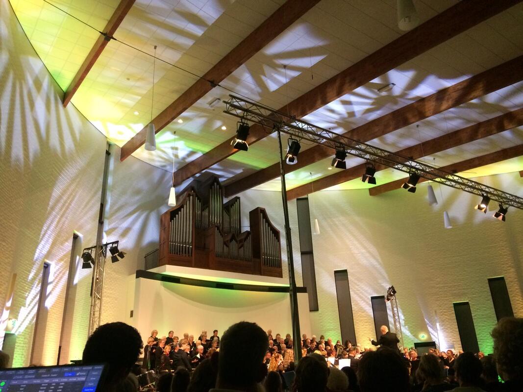 Concertkoor op het podium onder warm geel licht in een zaal met houten balken en een orgel op de achtergrond.