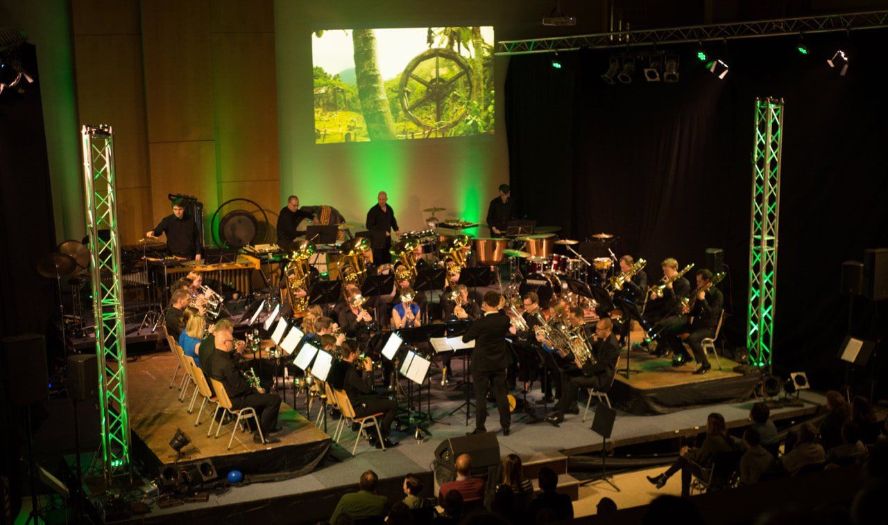 Orkest treedt op op een podium met warme verlichting en een groene geprojecteerde achtergrond.