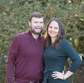 Two people smiling and standing together outdoors in front of greenery.