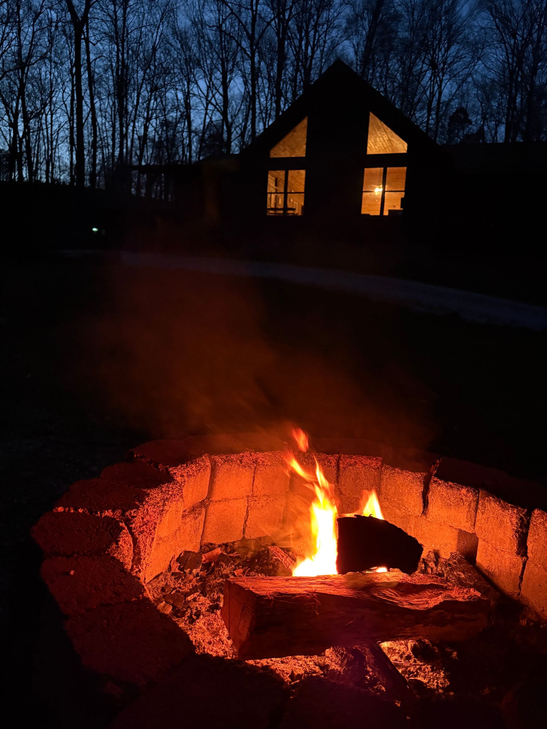 A backyard fire pit glows at dusk, with a lit cabin window and silhouetted trees in the background