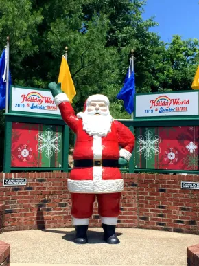 Santa statue waving outside a holiday world entrance, with festive signs and flags behind him