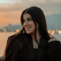 Young woman with long dark hair smiles, looking towards blurry city lights at sunset.