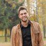 Man in a tan jacket and black shirt smiling outdoors with trees in the background.