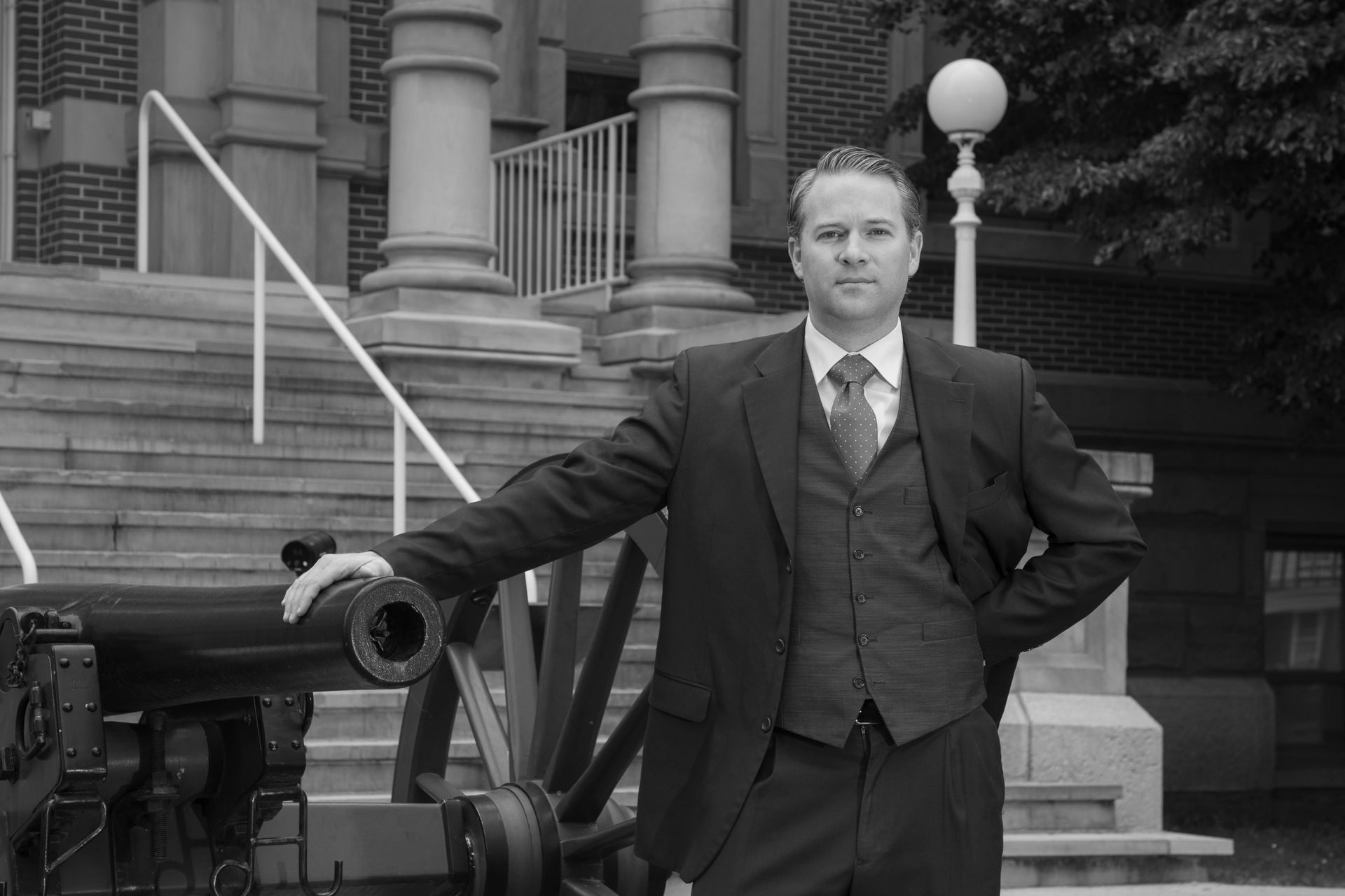 Man in suit, standing next to cannon in front of building with columns.
