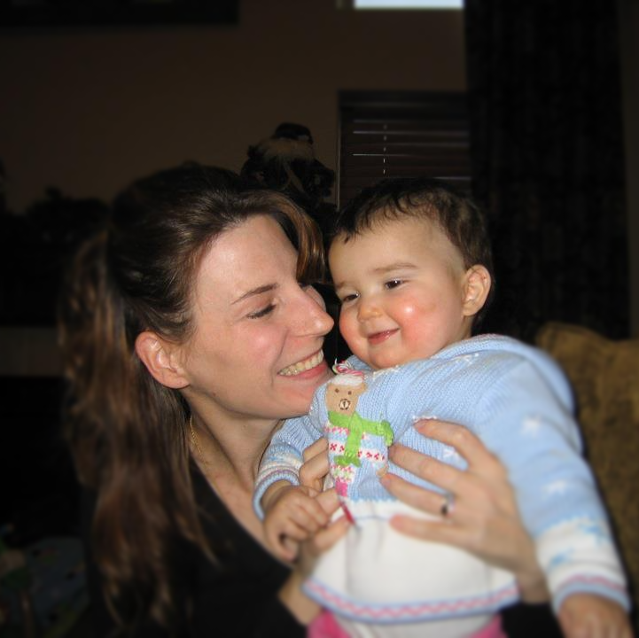 Woman with long brown hair smiles at baby in a blue sweater, indoors.