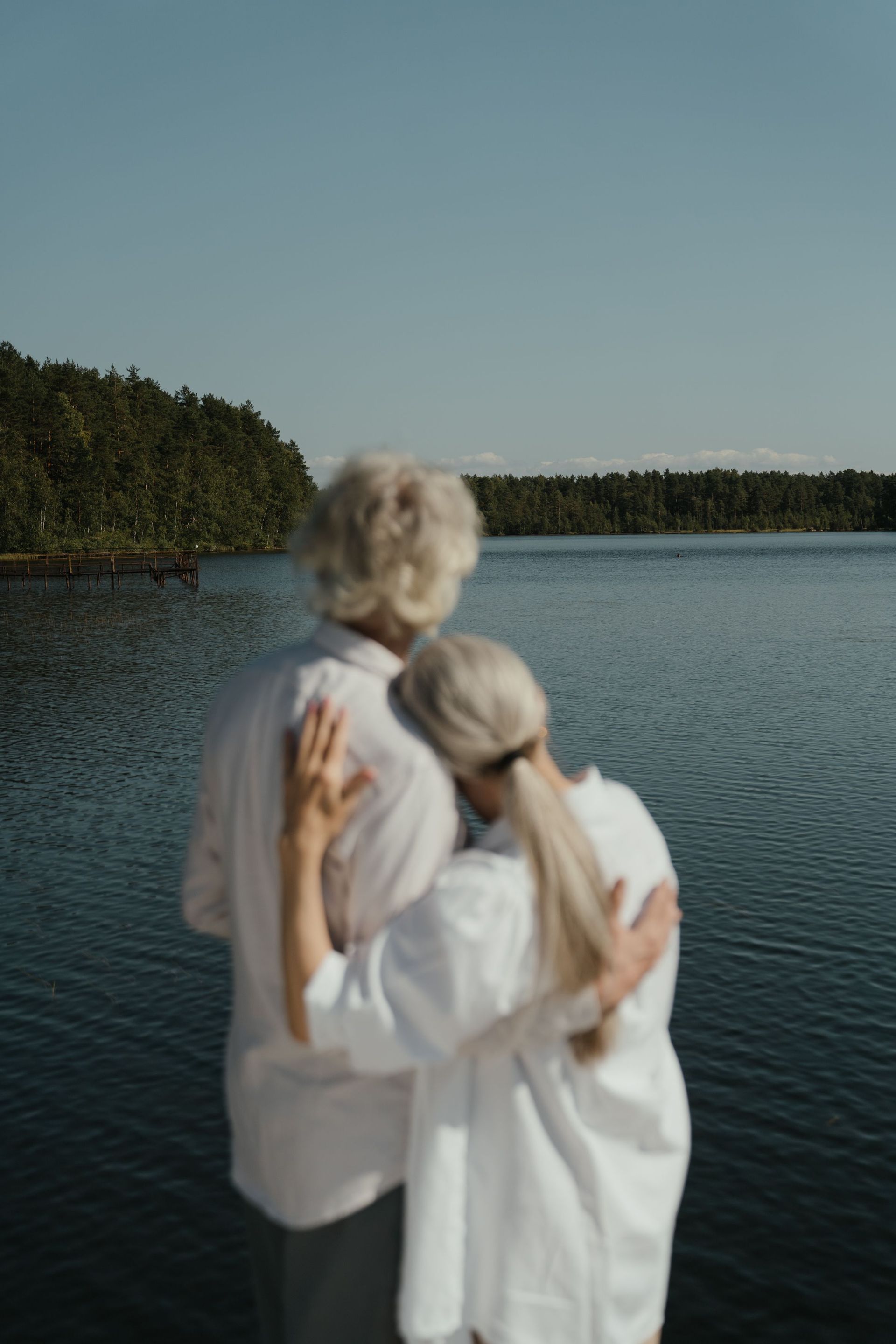 Two people in white shirts stand by a calm lake, facing the water and forested shore.