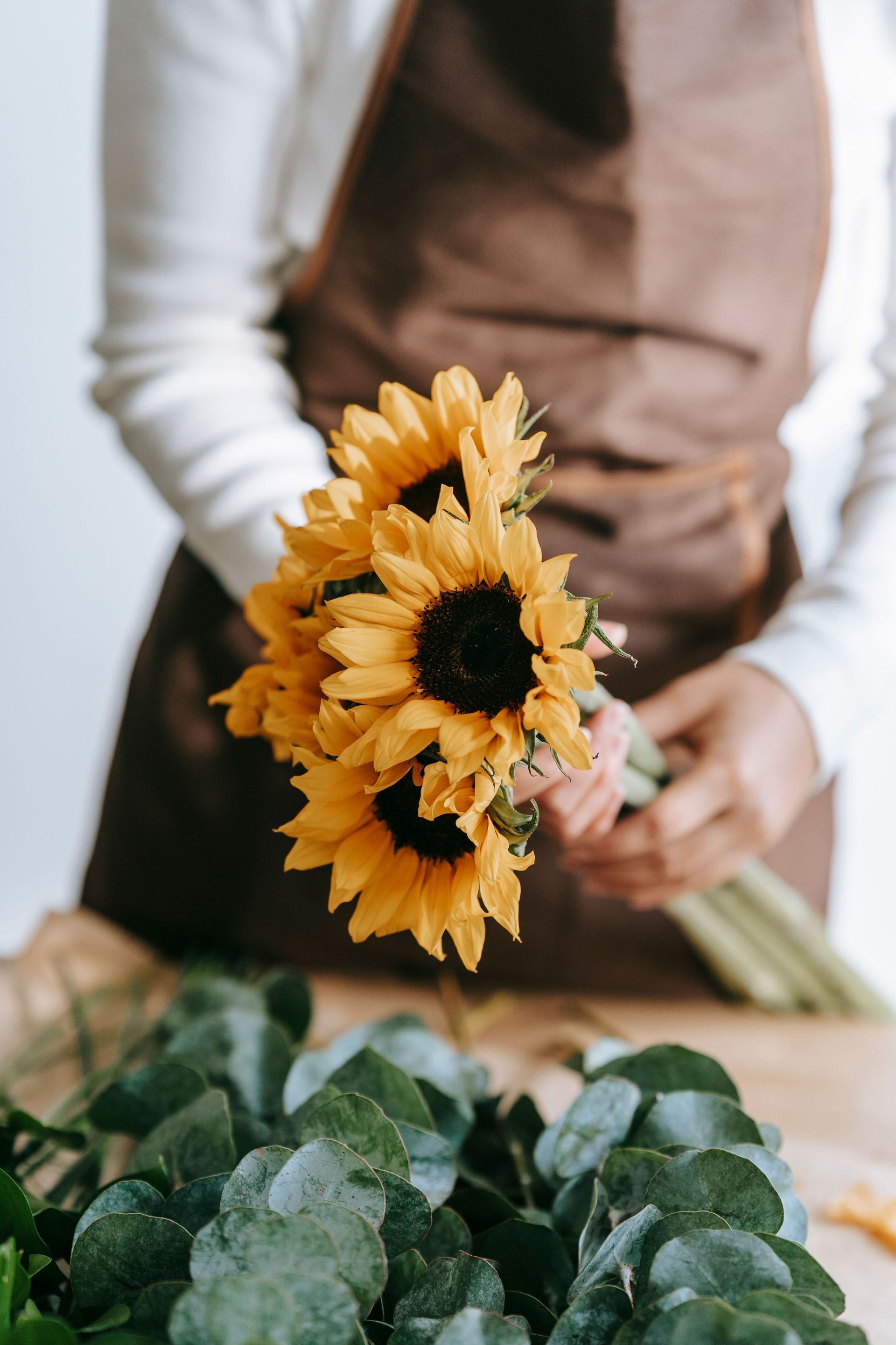 Person holding a bouquet of yellow sunflowers above a table with green leaves