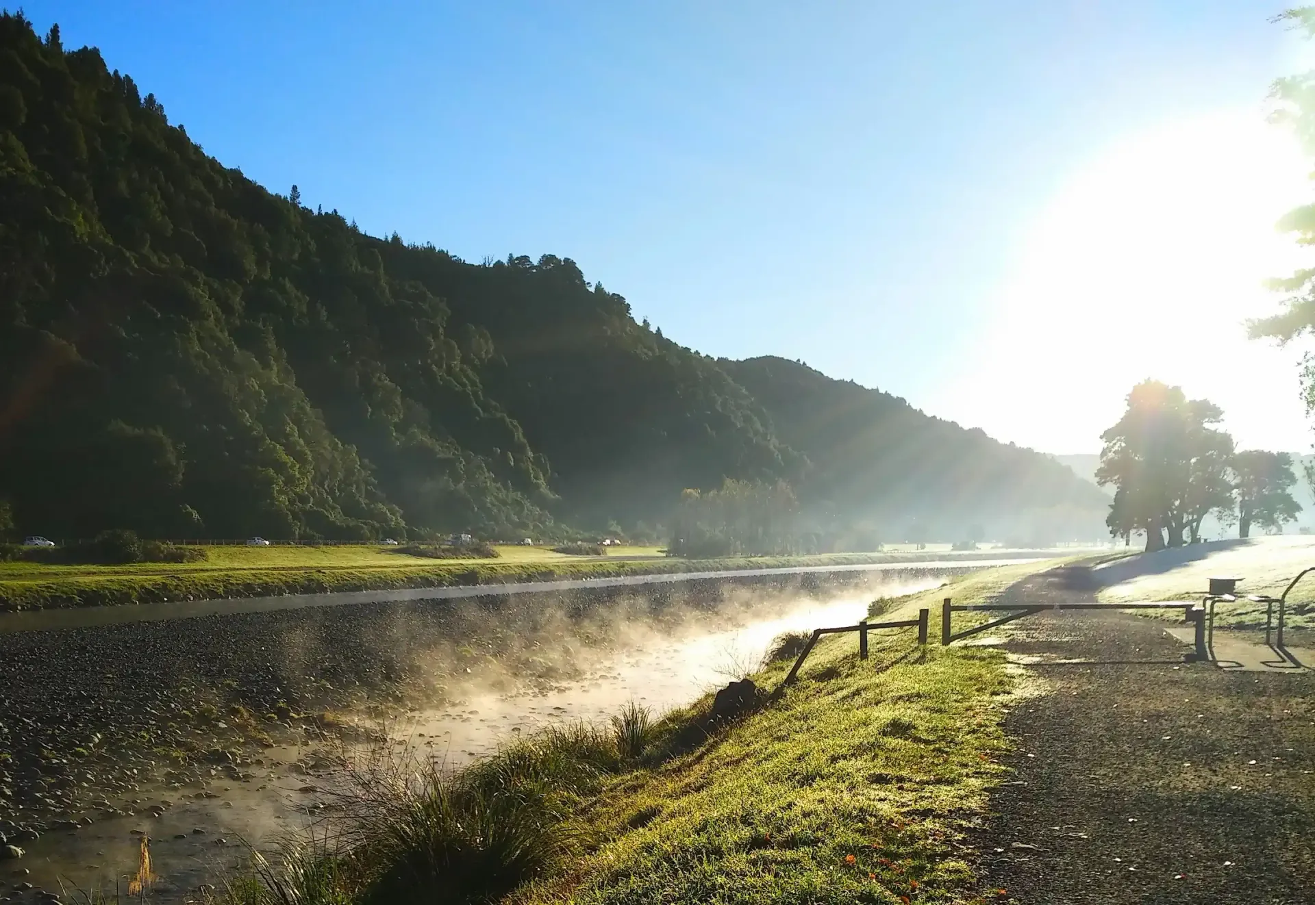 Sunlight over misty river with green banks and forested mountain.