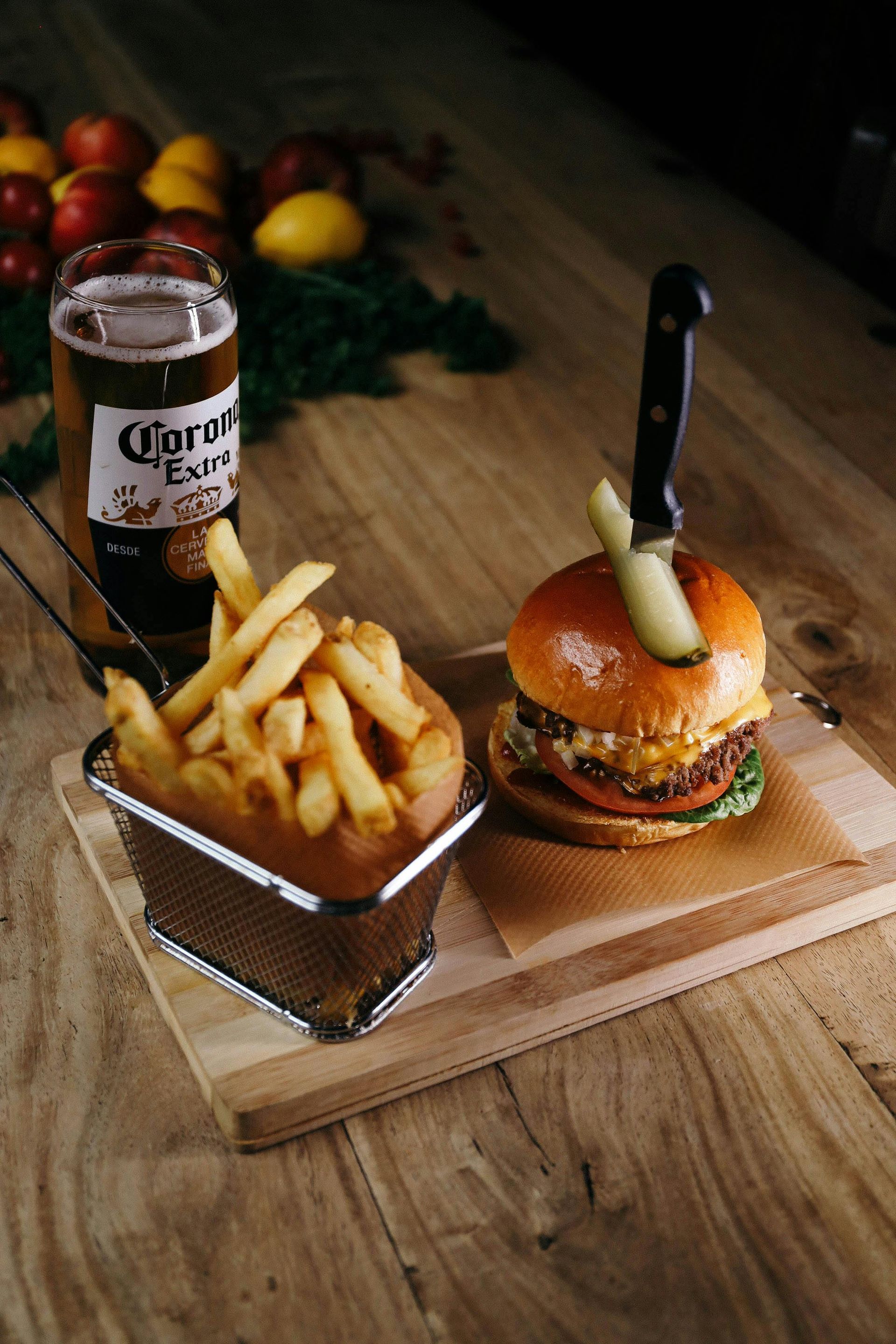 Burger with pickle skewer, fries, beer, and fruits on a wooden table.