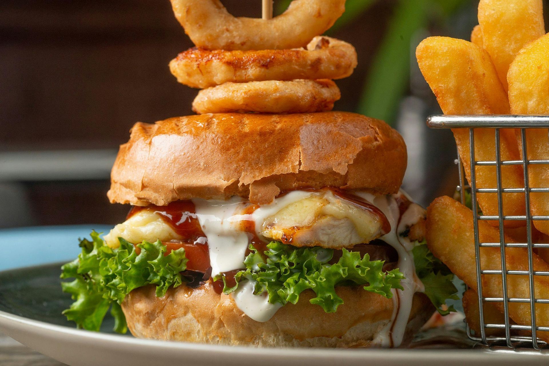 Burger with onion rings on top, served with fries in a metal basket on a plate.