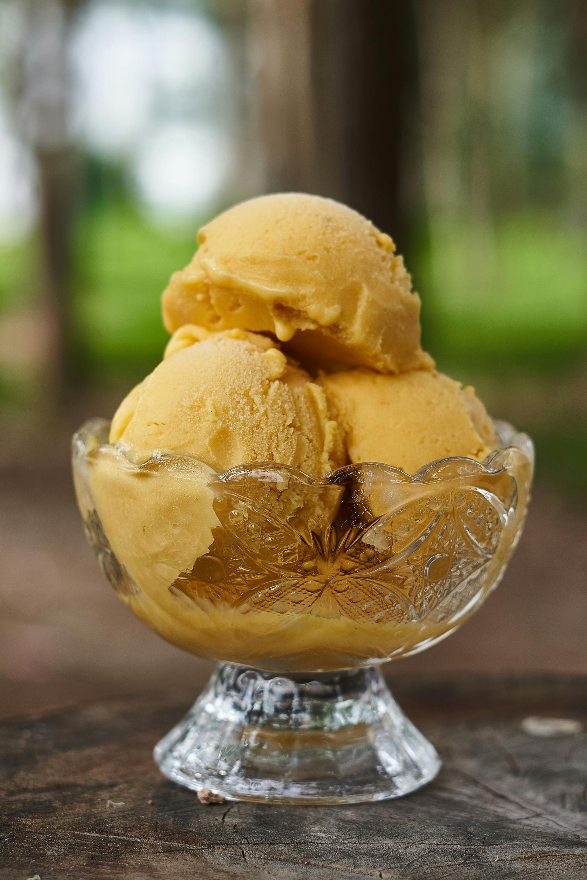 Scoops of yellow-orange ice cream in a clear glass dessert bowl, set outdoors.