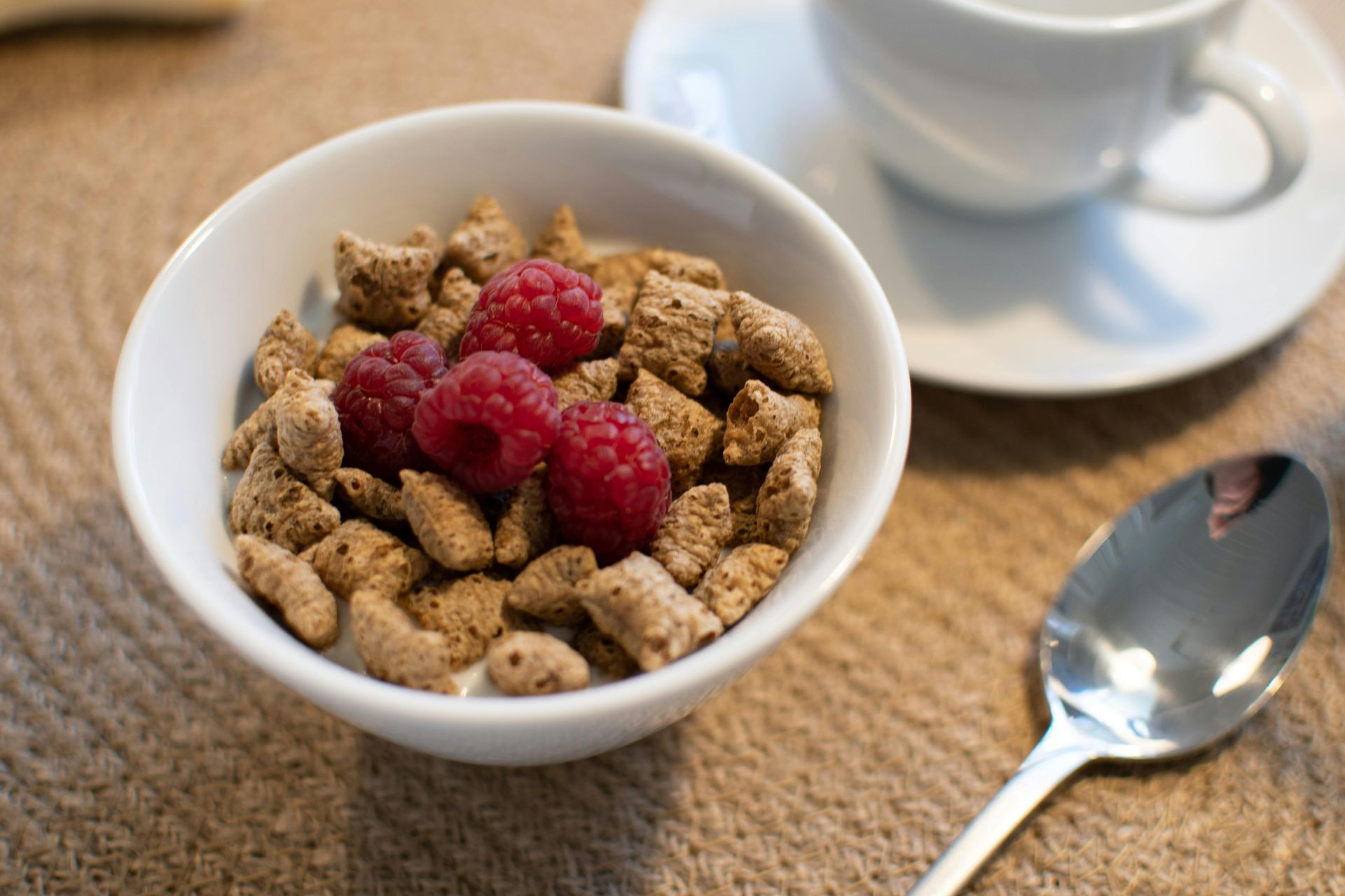 Bowl of cereal with raspberries, spoon and coffee cup on a textured surface.