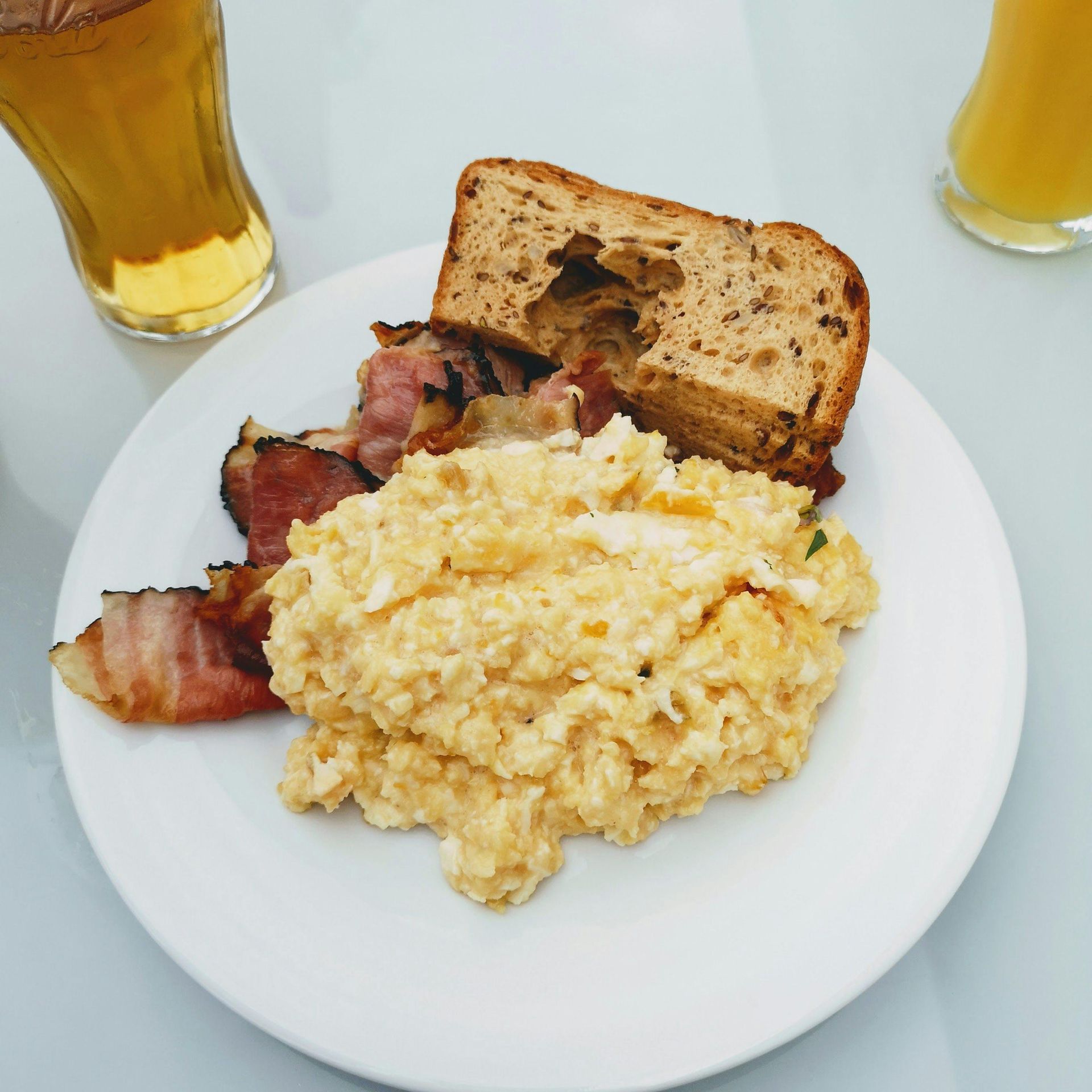 Scrambled eggs, bacon, and bread on a plate, with drinks in the background.