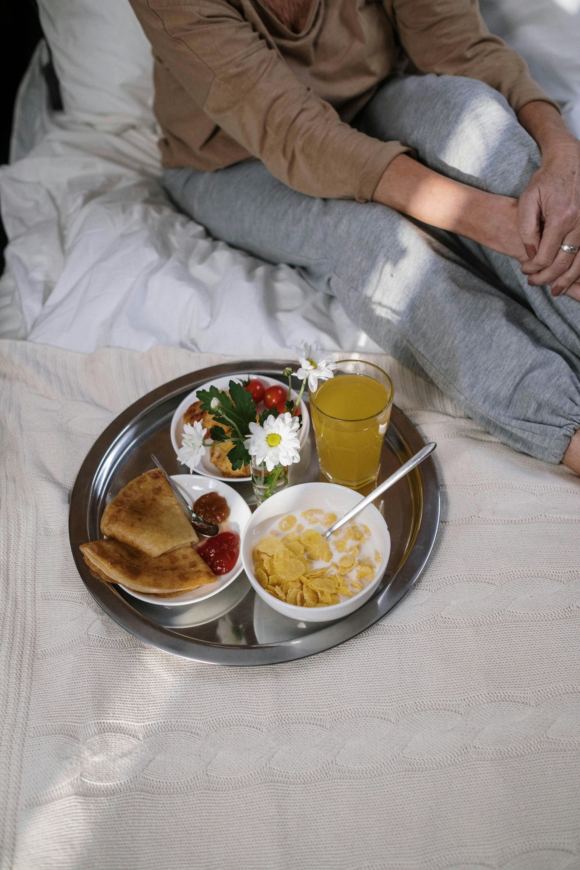 Person on bed with breakfast tray: toast, yogurt, cereal, orange juice.