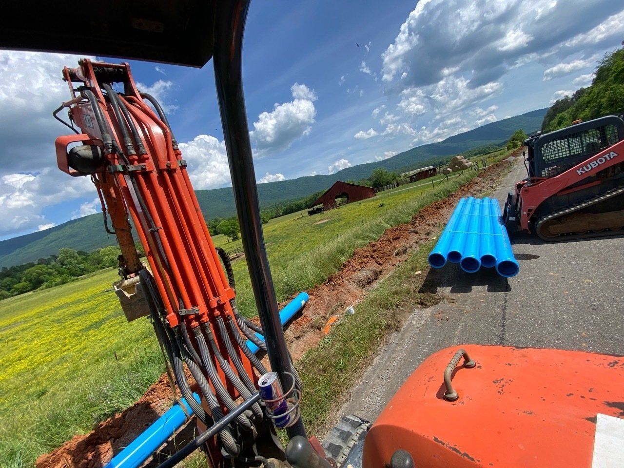 Orange farm machinery beside blue PVC pipes on a rural roadside under a cloudy sky