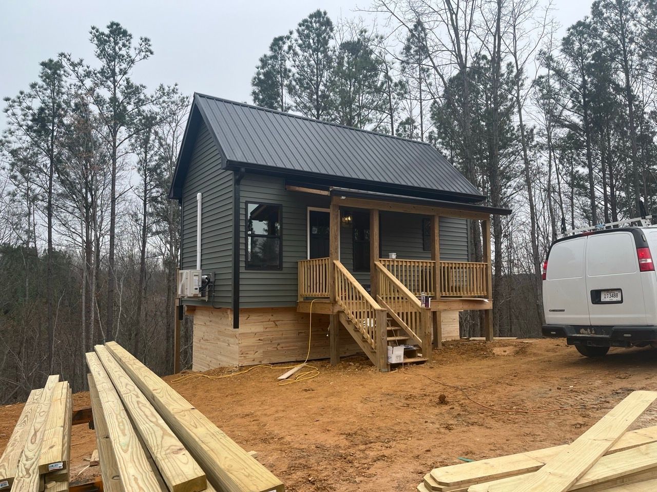 Small cabin under construction in a wooded area, with lumber stacks and a white van nearby.