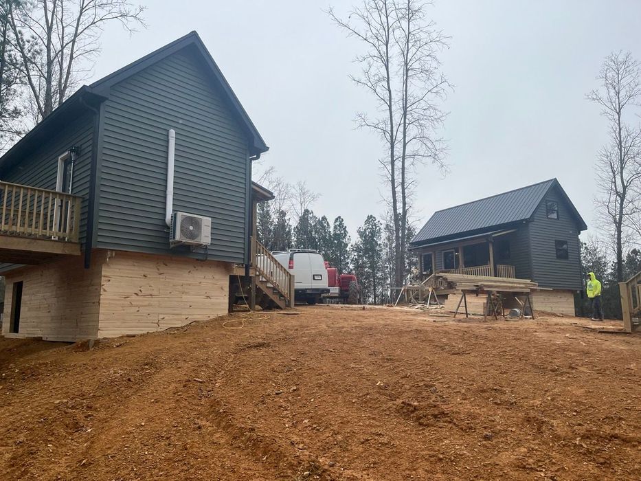 Two dark cabins on a muddy hillside with a white truck and tall trees in foggy weather